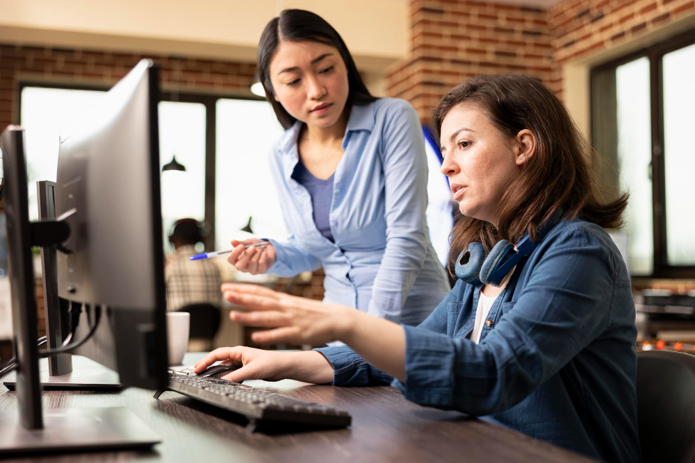 Photo of a woman in a blue shirt pointing at a computer screen for a colleague.