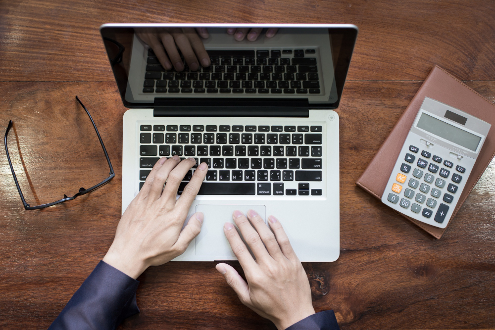 Close-up photo of hands typing on a laptop on a wooden table.