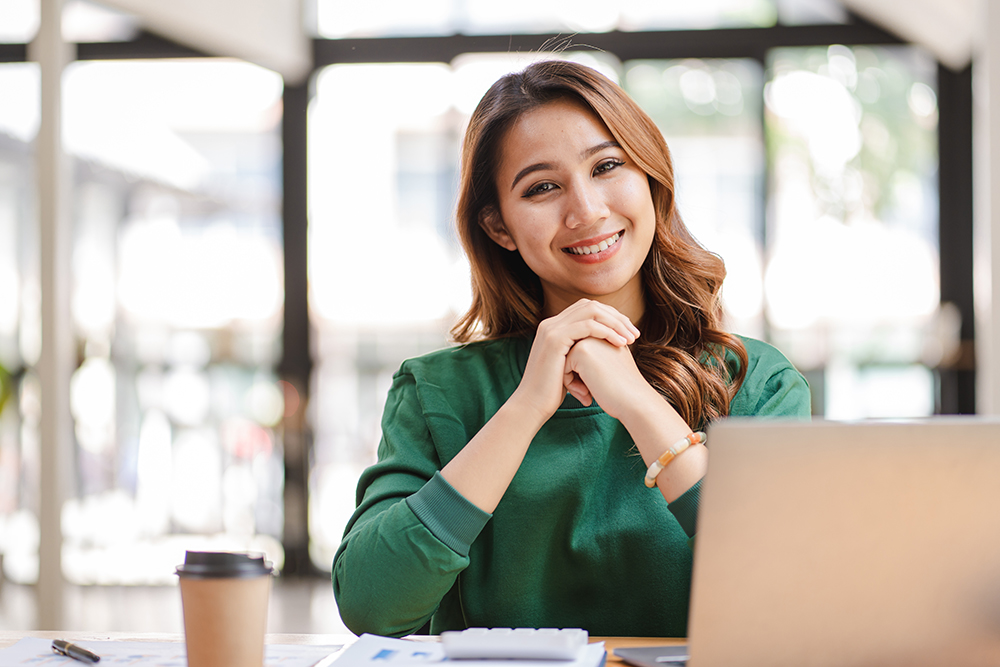 A professional Altselino executive virtual assistant smiling while sitting at a desk with a laptop and coffee in a modern office.