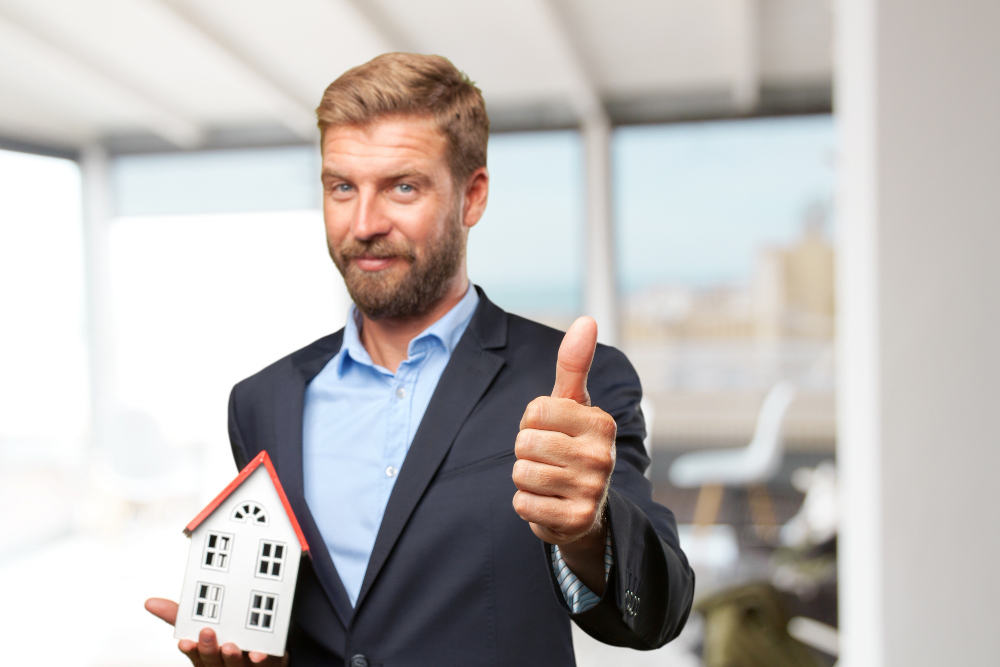 A professional man holding a small house model and giving a thumbs up, representing Altselino real estate VAs
