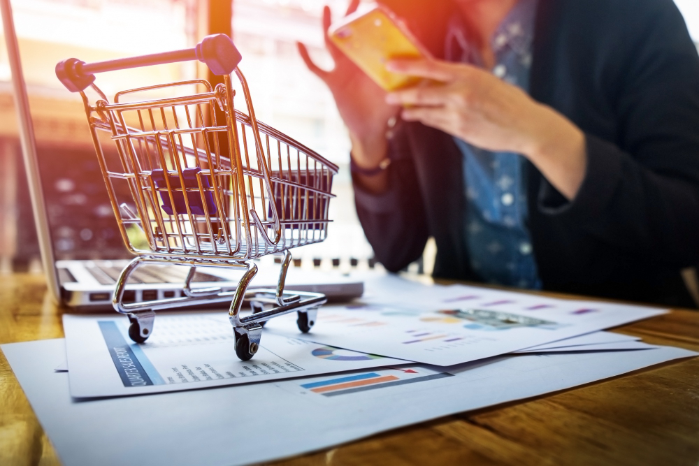 A miniature shopping cart on a desk with financial charts and a person using a smartphone.